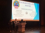 Two women stand on stage behind podium with a PowerPoint slide in the background