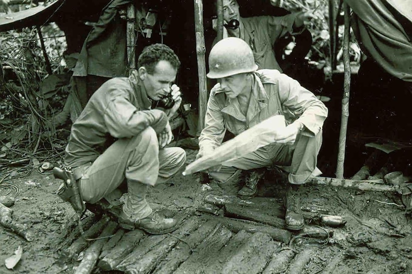 Two soldiers huddle together beside a mud hut, both looking at a map. One is pointing at the map while the other has a phone to his ear.