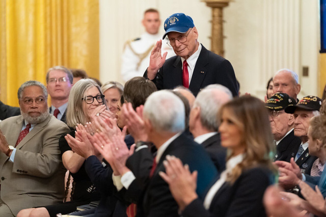 Man stands and salutes as a group of people sit surrounding him and clap.