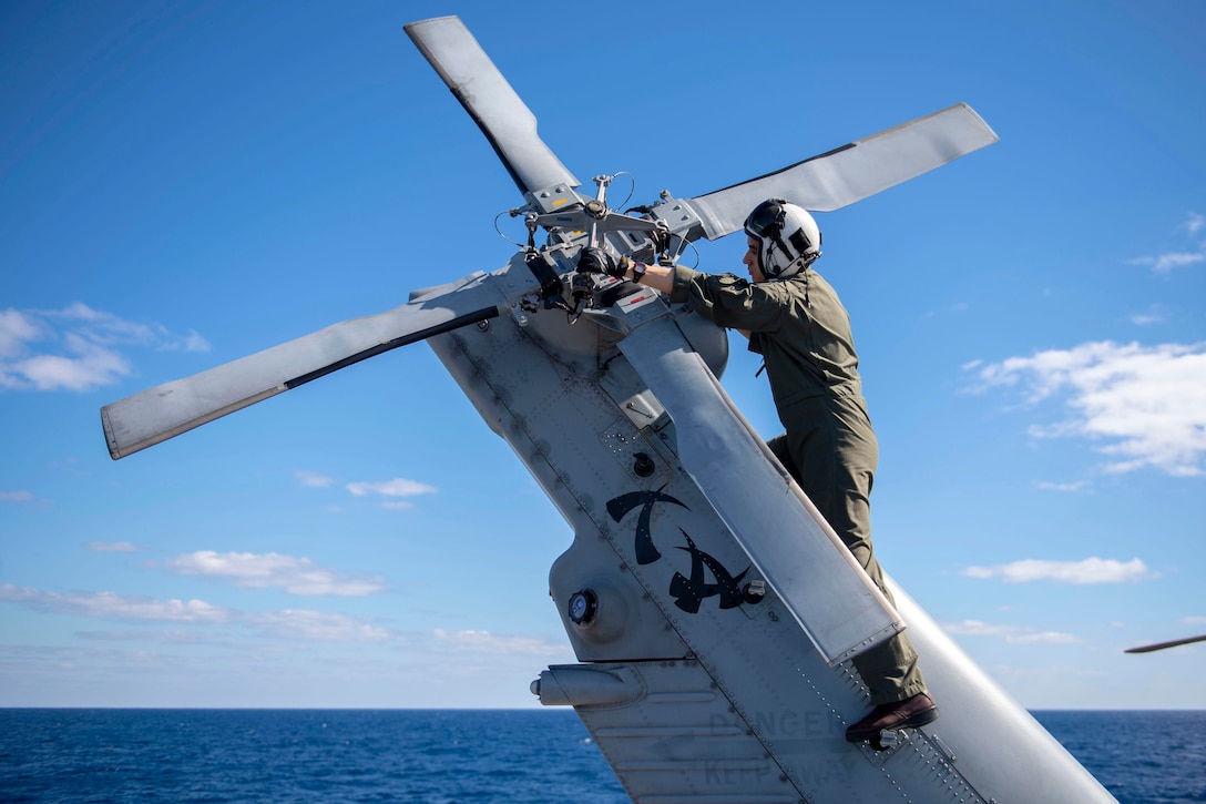 A sailor stands on the tail of a helicopter while checking the rotor.