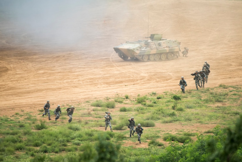 Troops and tracked vehicle move across terrain.