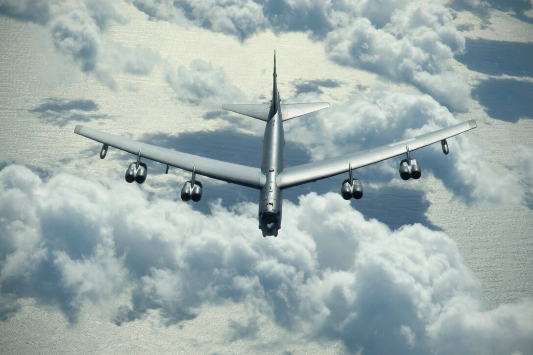 A large aircraft flies above clouds and the ocean.