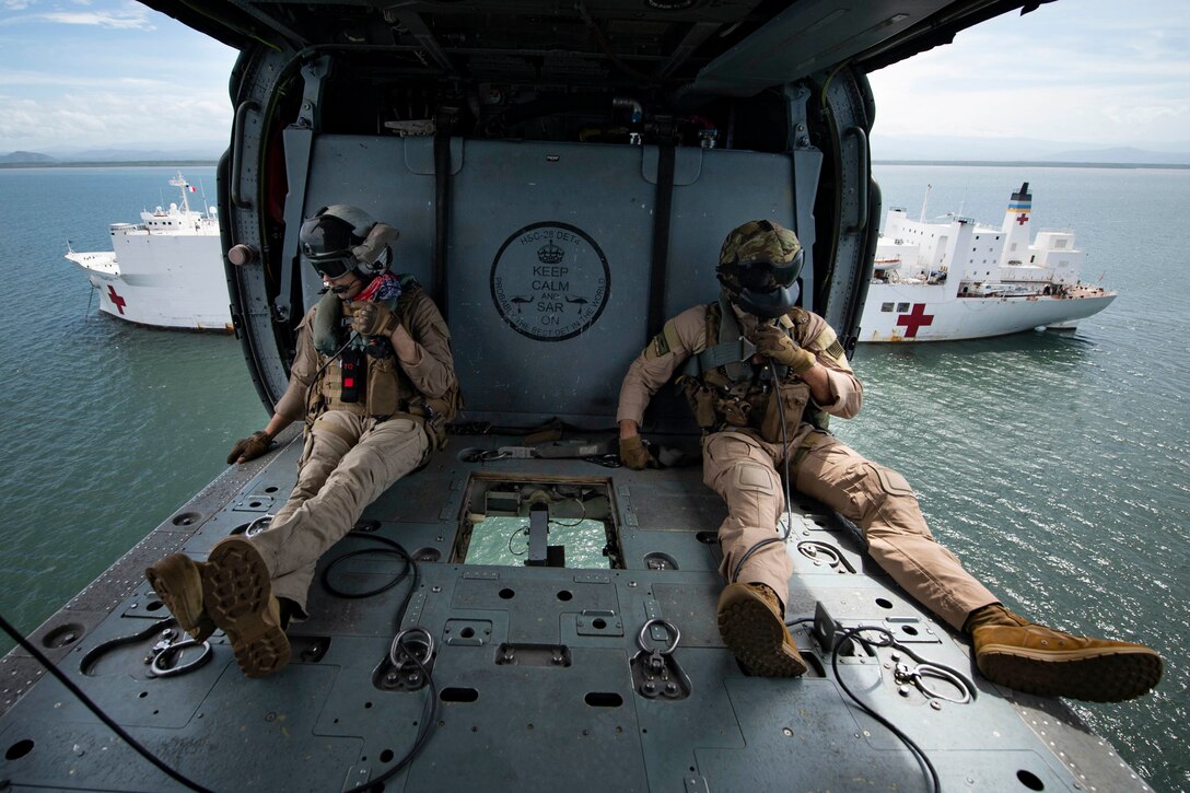 Two men sit in an open helicopter as it flies above the water with a hospital ship behind it.