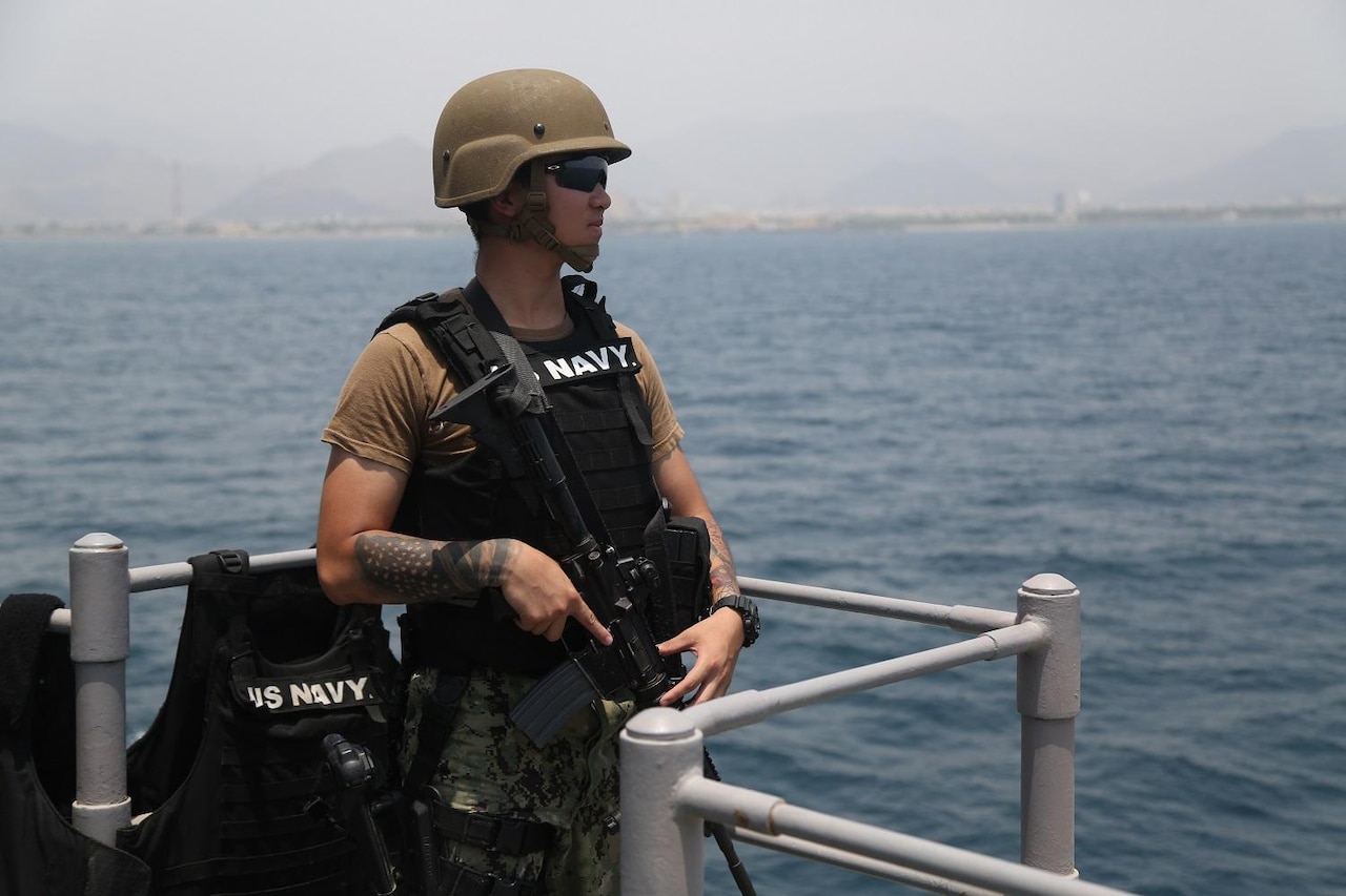 Sailor wearing a black vest and a green helmet stands watch on a ship.