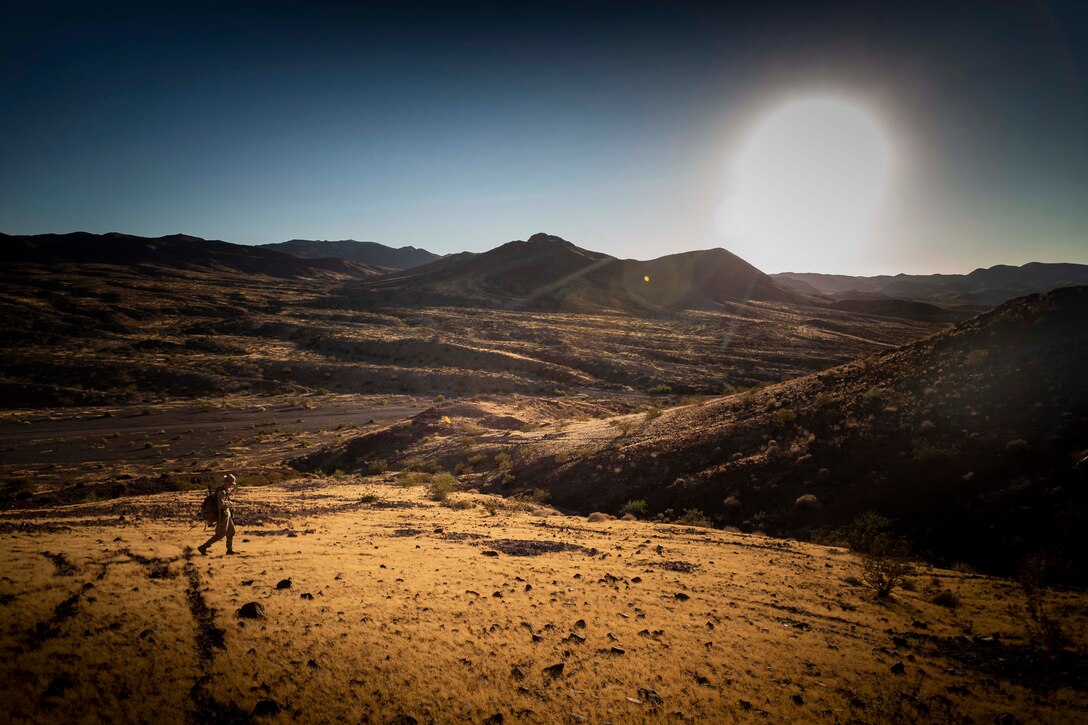 A Marine walks through a desert terrain with mountains in the background and the sun shining high in the sky.