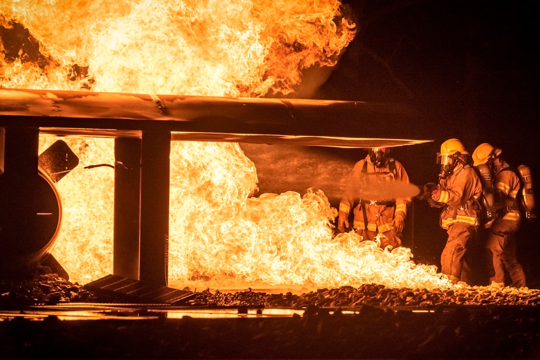 Three people spray a large airplane that is on fire with water.