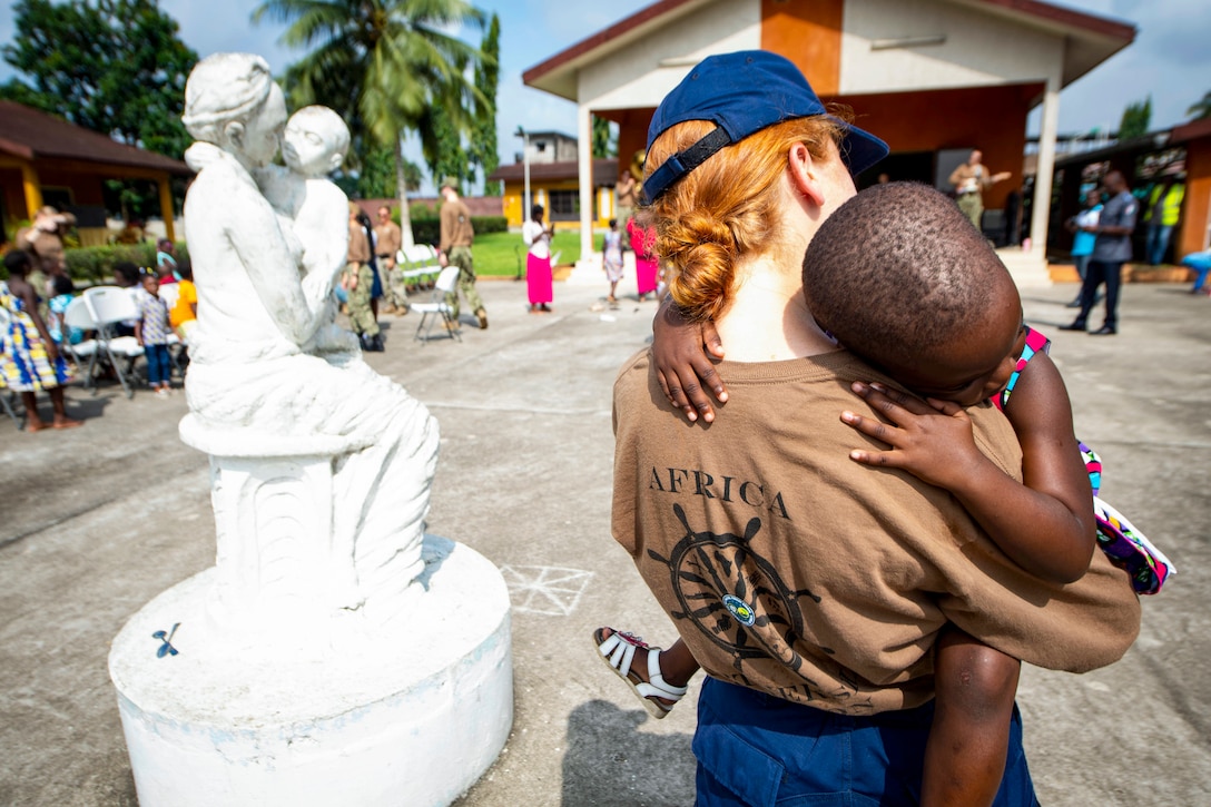 A Coast Guardsman holds a child while standing next to a statue of someone holding a child.