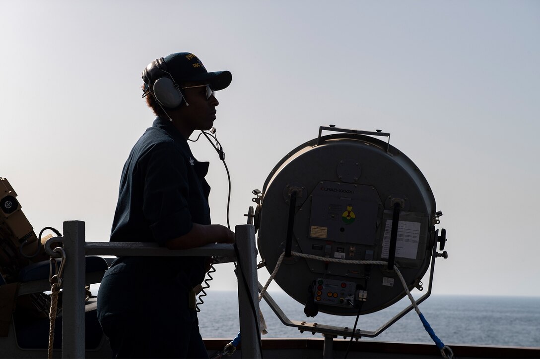 A single individual in silhouette wears earphones and looks out over the ocean. In front of her is a large circular spotlight.