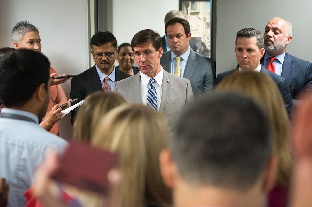 In a small room, several dozen individuals gather around a man in a light gray suit.