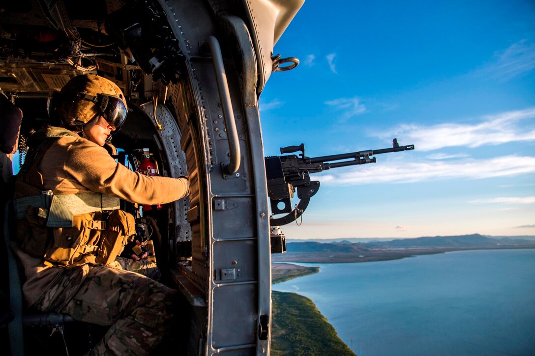 A sailor flies in a helicopter pointing a weapon toward the sky.