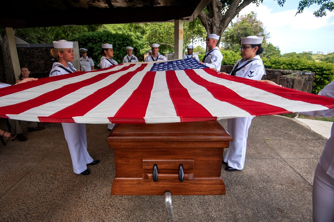 Sailors hold a flag over a casket.