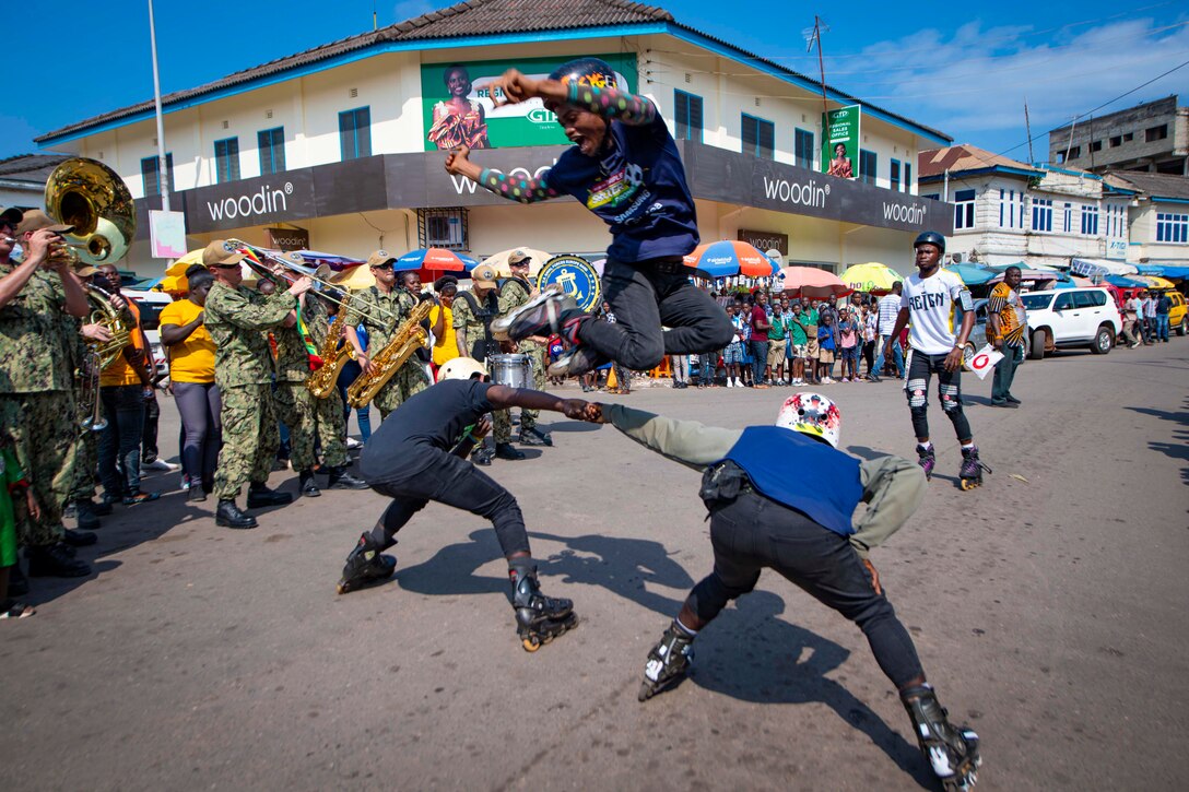 Three people wearing roller skates perform in the street surround by a group of people, some playing instruments.