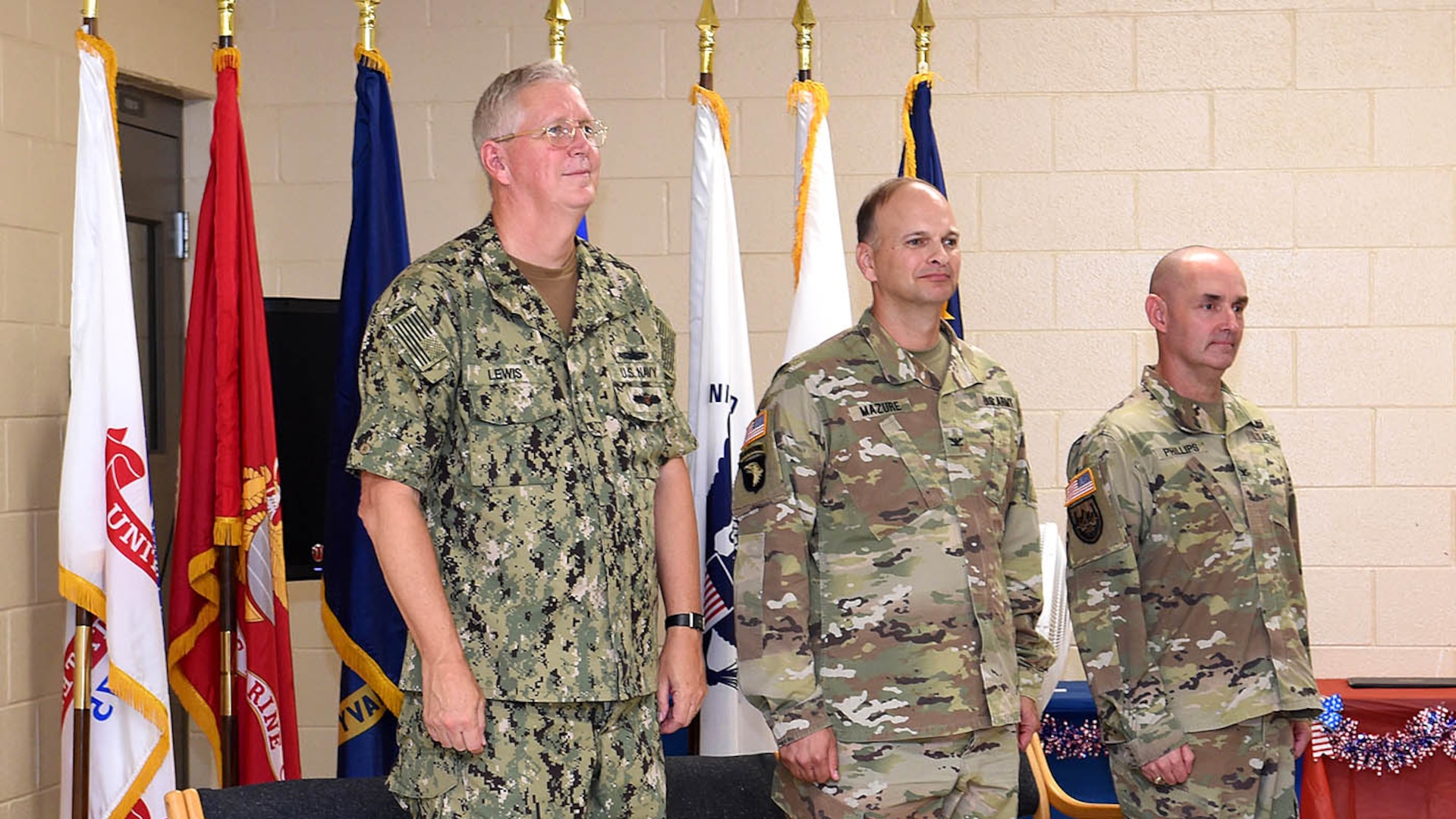 Three commanders standing at attention in front of U.S. and service flags.