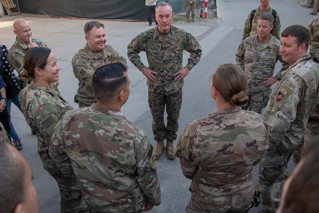Service members in camouflage uniforms talk while standing in a circle.