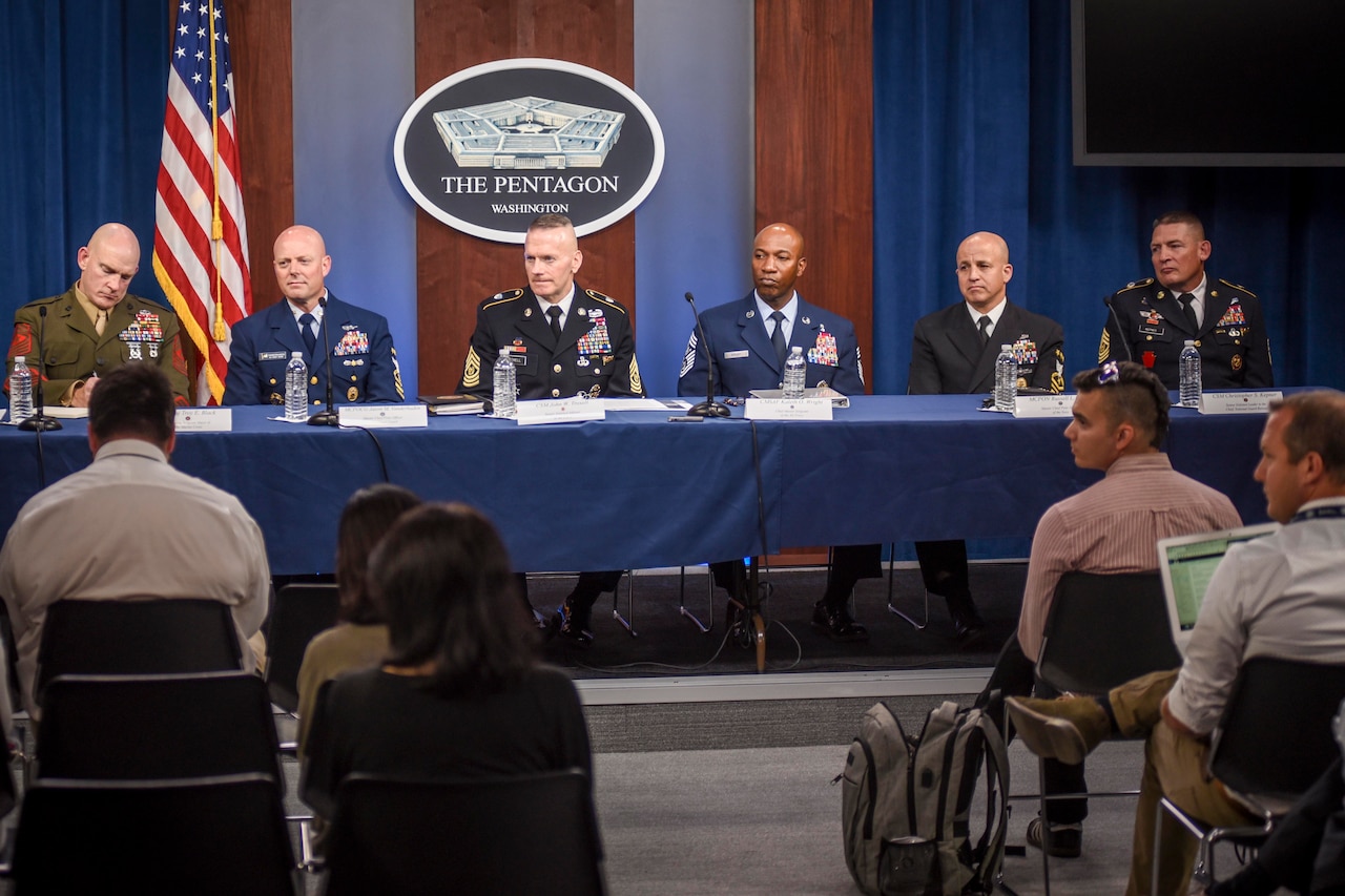 Six men in military uniforms are seated at a long table. In the background is an American flag, and a sign bearing an image of the Pentagon.”