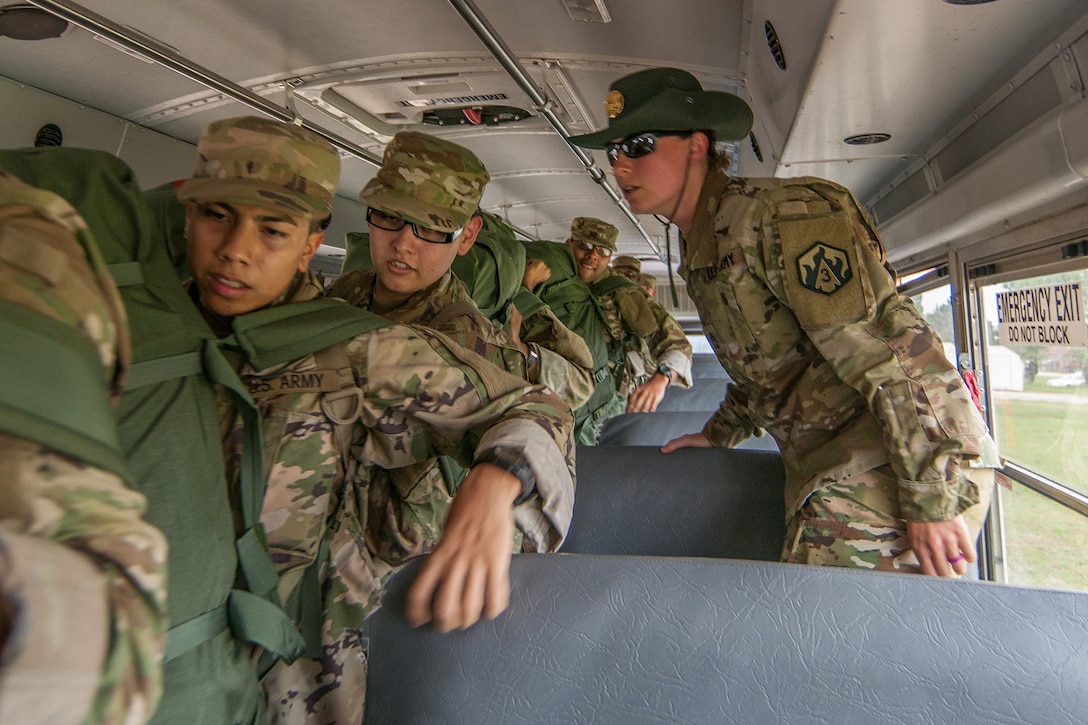 Young individuals in military uniforms and carrying large duffle bags are lined up in the aisle of a school bus, preparing to exit. Another uniformed service member wearing a “campaign hat” stands nearby.