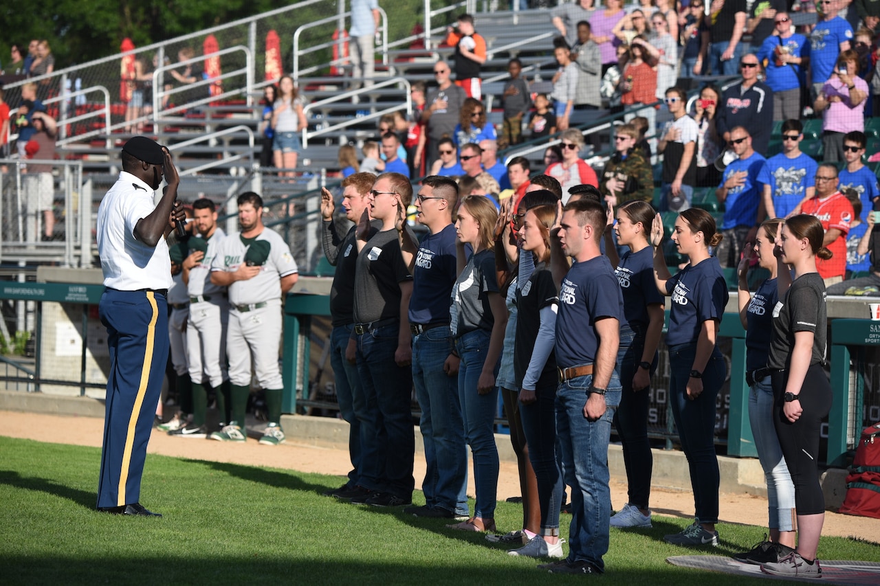 More than a dozen young individuals in civilian clothing stand in formation with their right hand raised above their heads. In front of them, a uniformed military officer also raises his hand and administers an oath.