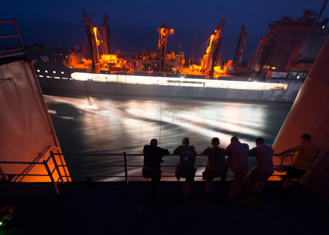 Troops look out at a ship from the deck of another at night.