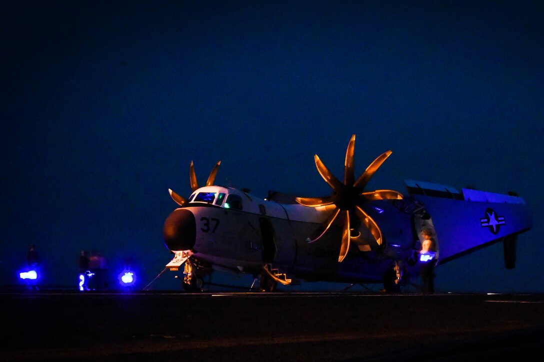 An aircraft sits bathed in blue and yellow lights on a ship's deck.