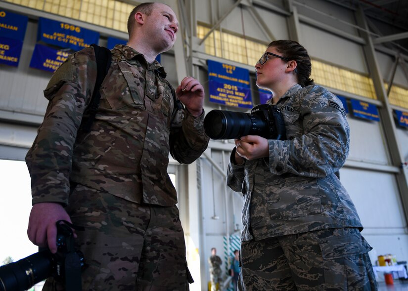 Senior Airman Cody Miller, left, a photojournalist assigned to the Joint Base Charleston Public Affairs Office, explains public affairs responsibilities to Senior Airman Fawne Maguire, an aerospace maintenance journeyman assigned to the 437th Aircraft Maintenance Squadron, before a change of command ceremony at Joint Base Charleston, S.C. July 12, 2019. Maguire said she is interested in retraining out of her current career field and into the public affairs, comptroller, personnel, manpower and aviation resource management career fields. She was able to learn more about the career fields by shadowing members of each job.