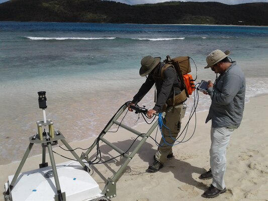 A man is pushing AGC equipment and another man is tapping a computer tablet attached to the AGC machine on Flamenco Beach