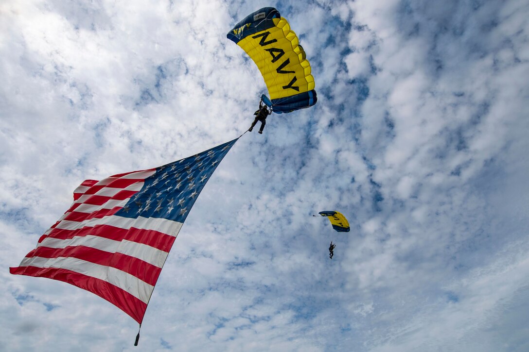 Two people wearing parachuted descend in the sky while one holds displays an American flag.