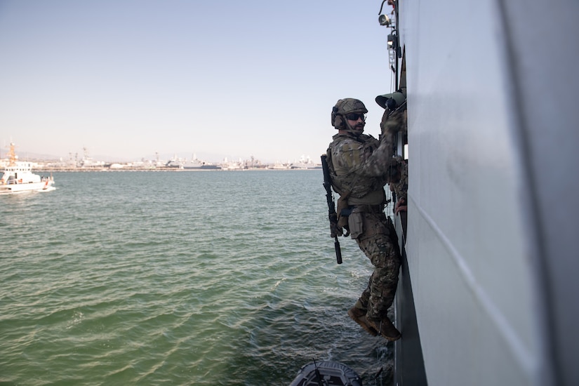 A service member climbs on the side of a ship in the water.