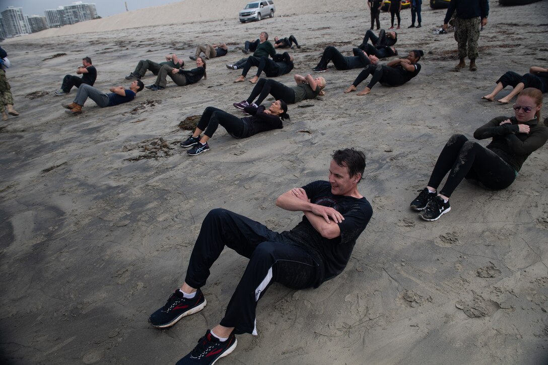 Civilians in black workout clothes do situps on sand.