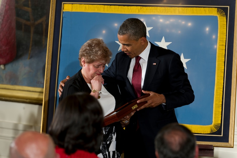 President Barack Obama has his hand around the shoulder of a woman who is holding and looking at a case with a Medal of Honor in it.