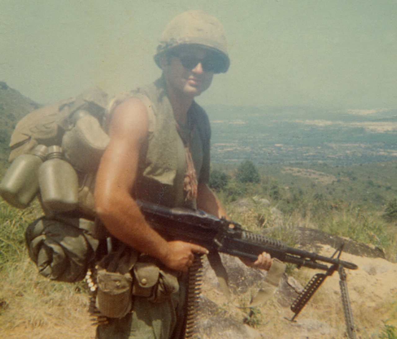 A sleeveless soldier wearing a helmet and carrying a backpack, lots of ammunition and several water bottles poses on a mountainside for a photo with his rifle.