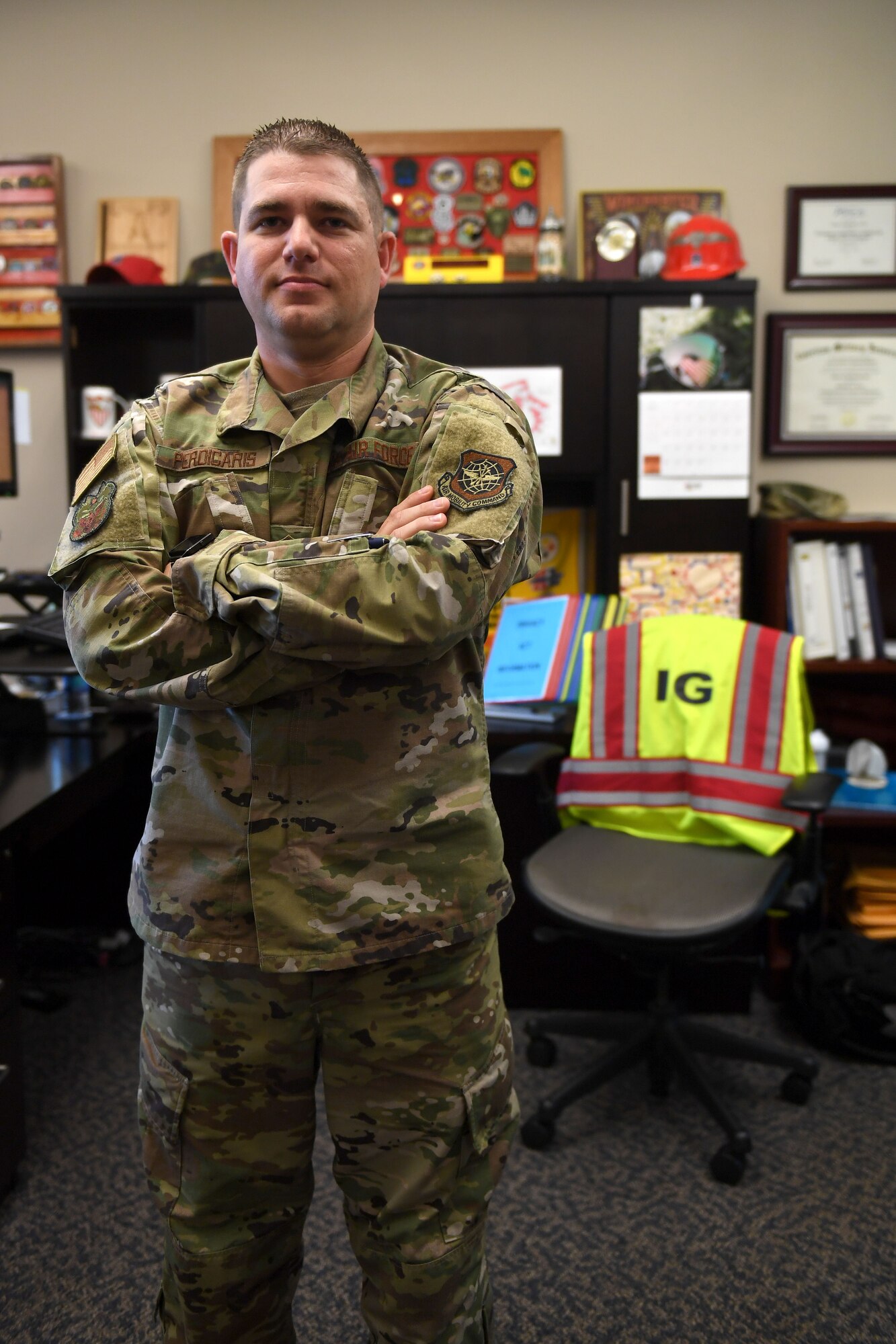 A man in a multi colored uniform stands with his arms folded in front of a desk.