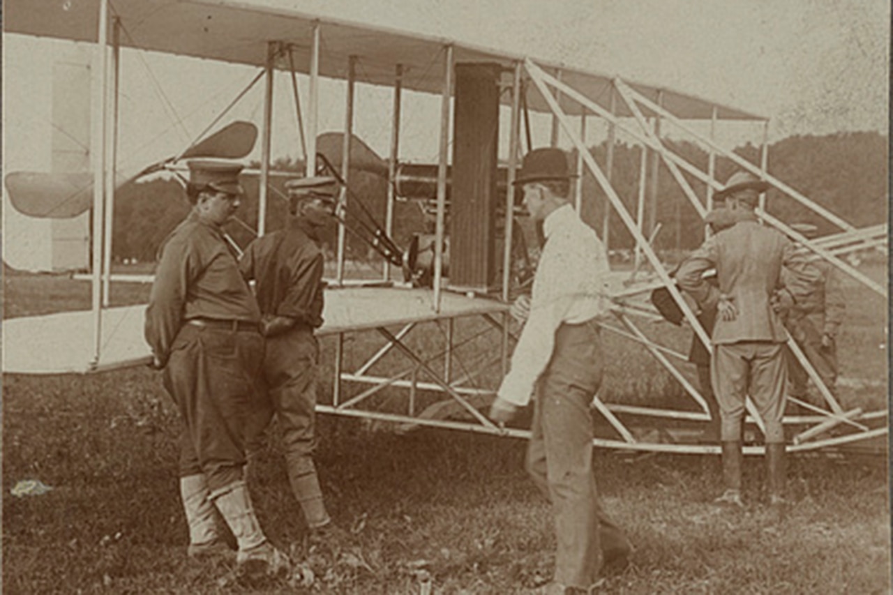 Six men in early 1900s garb stand in a field beside the earliest model of a military airplane.