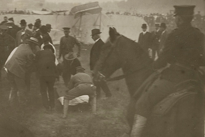 Several people work to put a man on a stretcher in a field with a crashed airplane on its side in the background. A soldier on a horse is in the foreground.