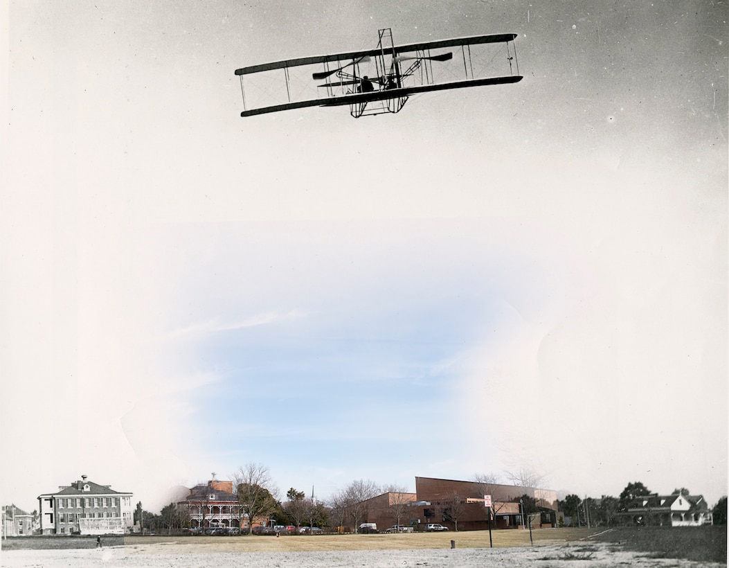 A Wright Military Flyer airplane flies in the skies above a field with buildings in the background.