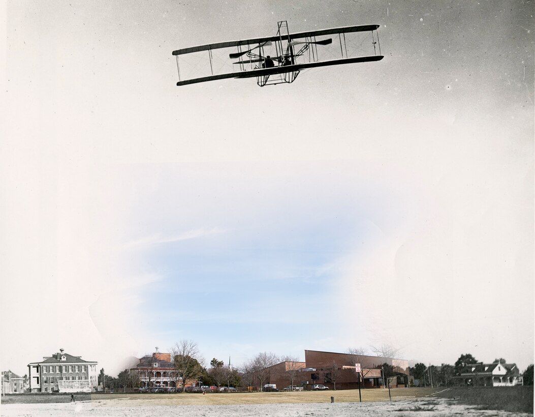 A Wright Military Flyer airplane flies in the skies above a field with buildings in the background.