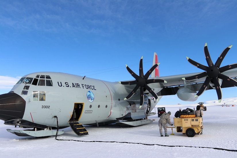 An Air Force LC-130 cargo plane with skis sits on a snow-covered runway. A line for fuel is attached to it, and two men work on a piece of equipment nearby.