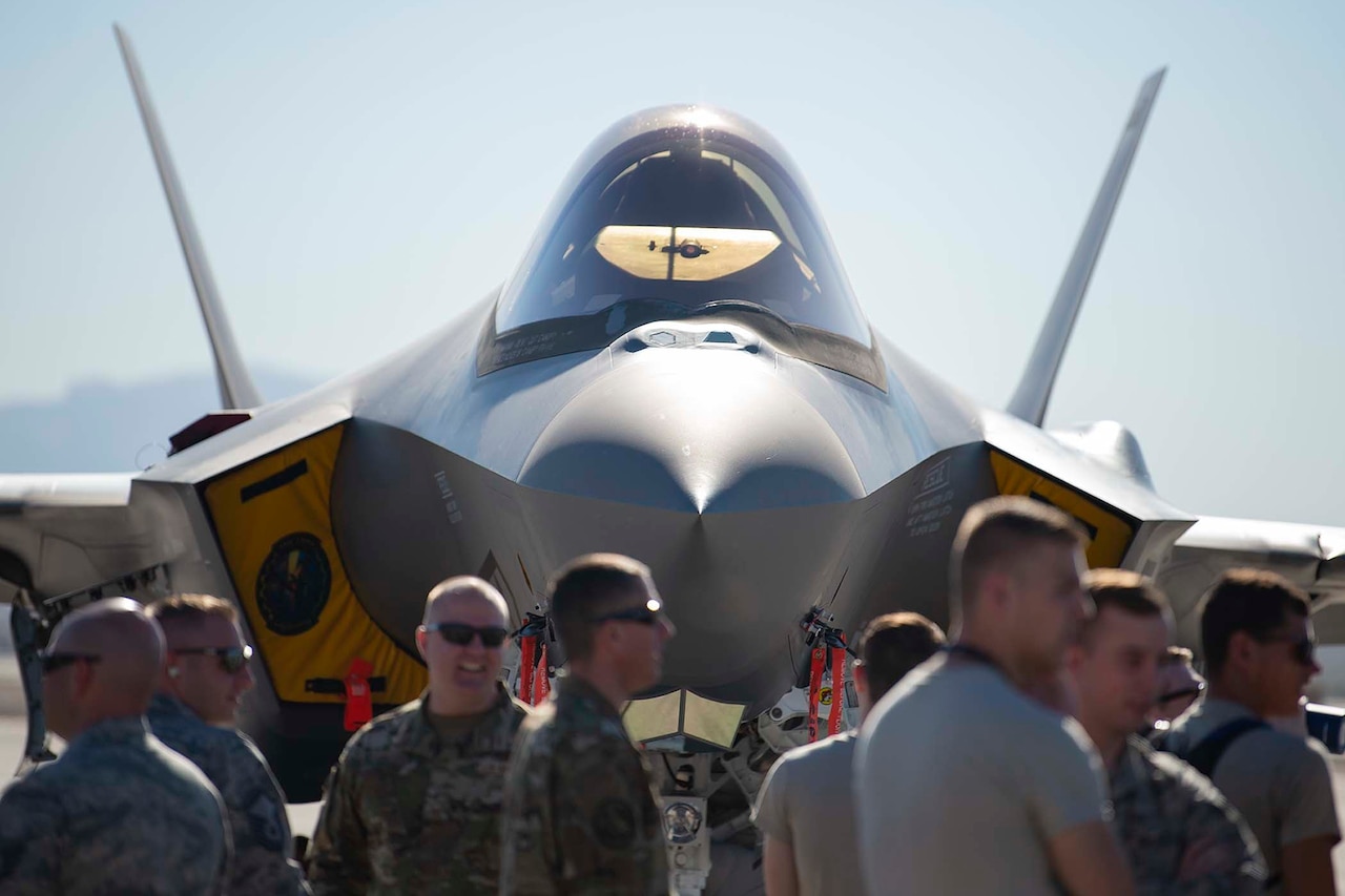 An F-35 Lightning II fighter jets sits on an airfield as several airmen mill around in front of it.