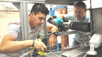 IMAGE: DAHLGREN, Va. (July 17, 2019) – U.S. Naval Academy Midshipman Max Shuman mounts a thermocouple assembly that houses spring fit thermocouples to a target plate during his internship at the Navy’s Laser Lethality and Development Facility which features an above-ground tunnel used for evaluating high energy laser weapon systems. Shuman’s mentor, Naval Surface Warfare Center Dahlgren Division scientist Chris Heflin, observes as the Midshipman prepares for a test shot accomplished with a flat top beam to ensure uniform heating across the sample.