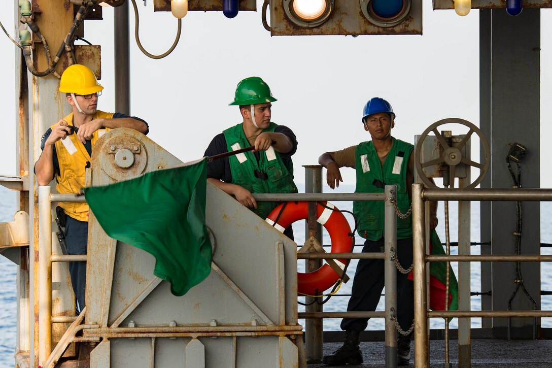 Three sailors stand on the deck of a ship; one holding a flag.