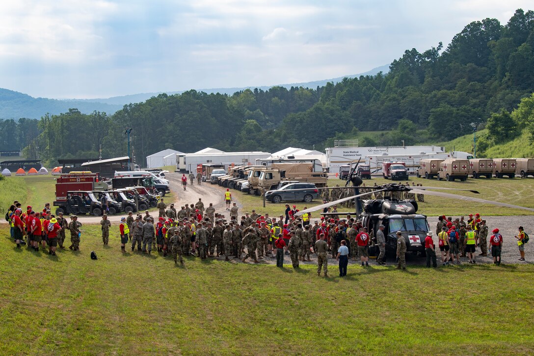 A small tent city sits in the center of a large clearing amidst a forested area.  Several dozen individuals, mostly military personnel, are gathered around a helicopter bearing a red cross symbol.  In the background are nearly a dozen military medical vehicles.