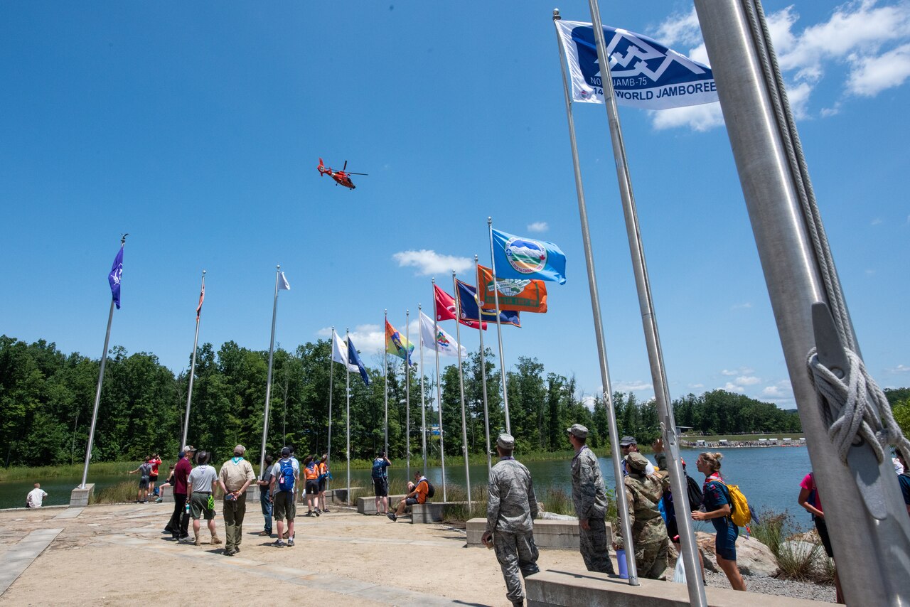 More than a dozen people look upwards to an orange helicopter hovering above a lake and tall trees.  They are surrounded by flagpoles.