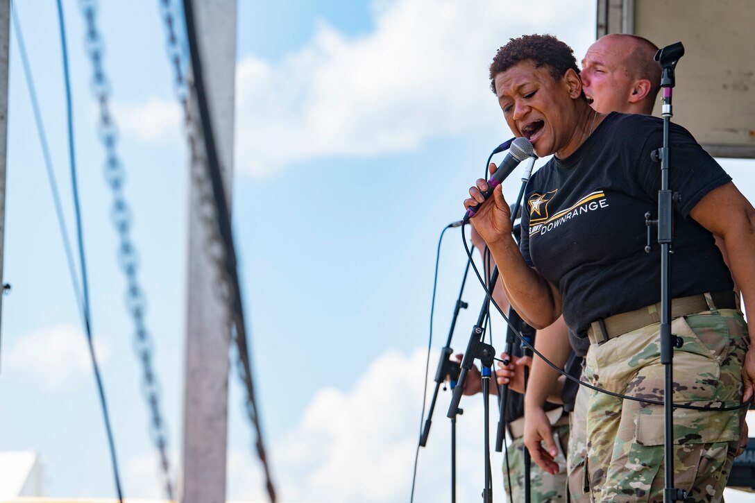 A musician wearing a black shirt and camouflage pants sings into a microphone.