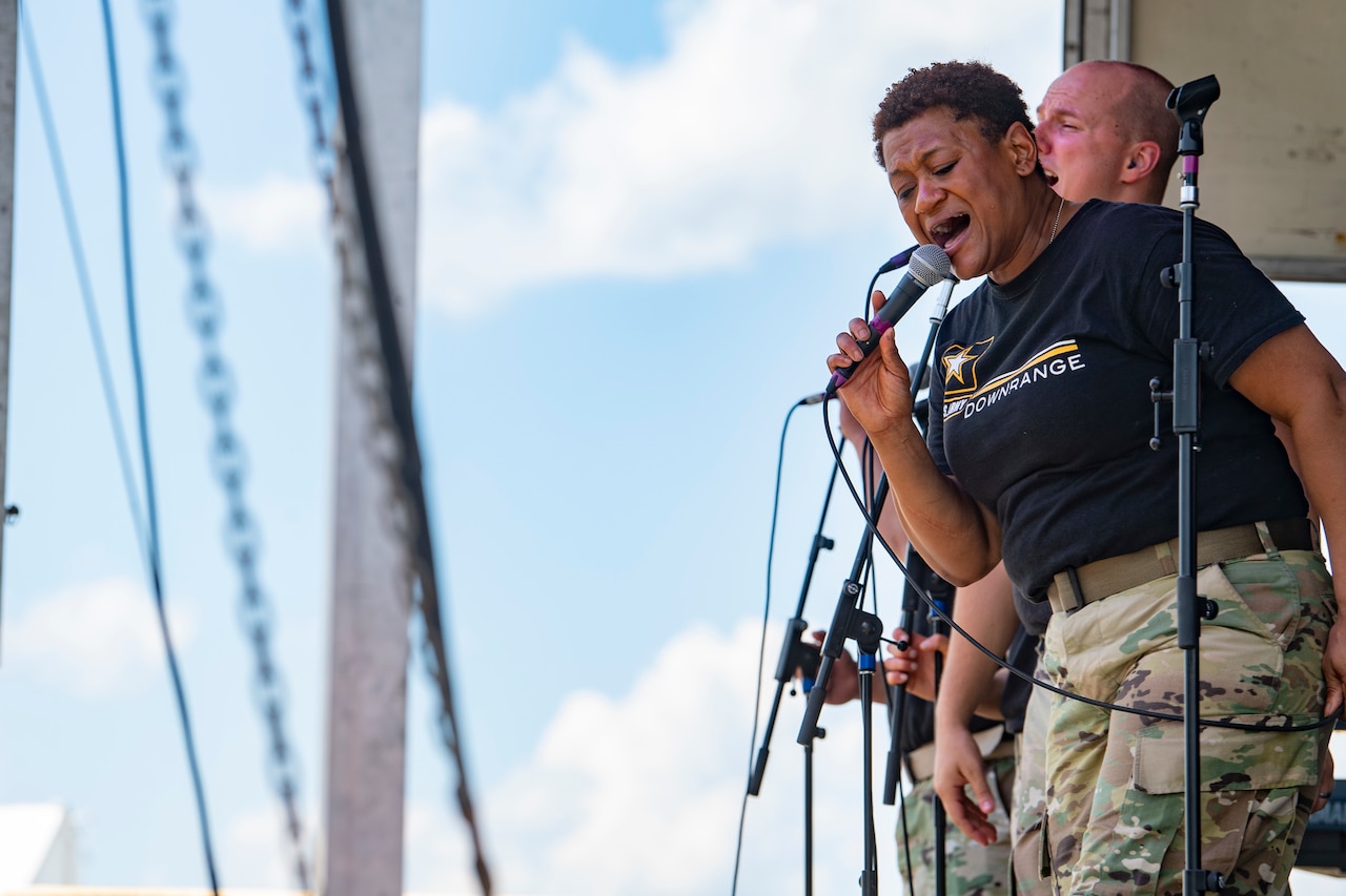 A musician wearing a black shirt and camouflage pants sings into a microphone.