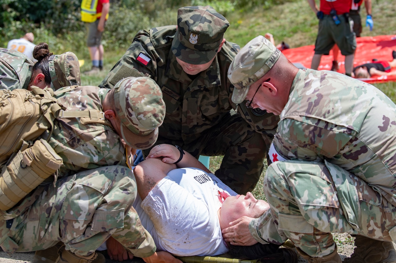 Multiple military personnel squat on the ground around a simulated medical patient.