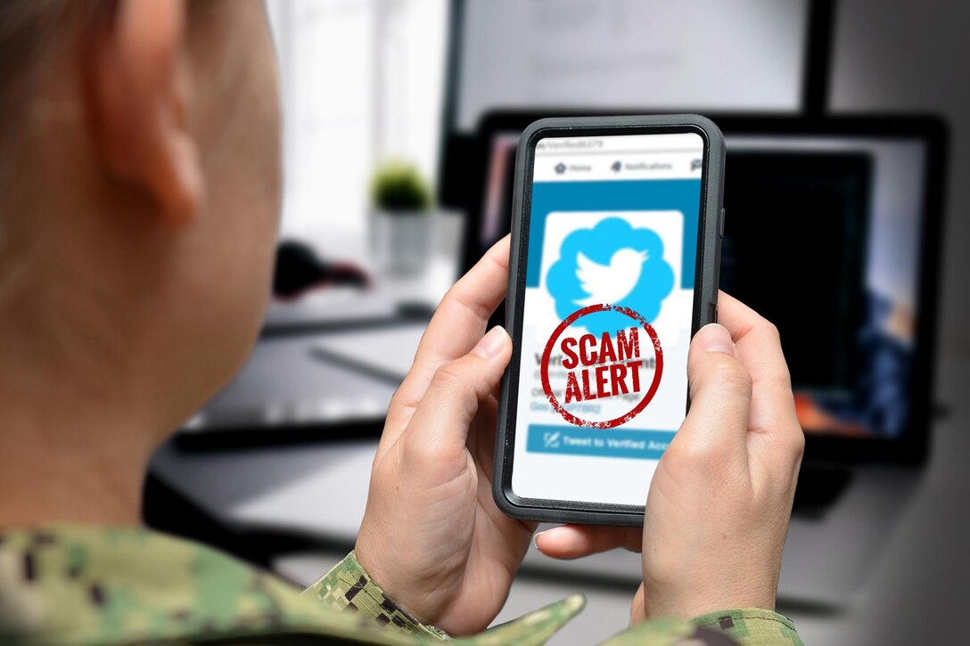 A Navy sailor holds up a smartphone, making visible a screen that shows a red circle with the words “scam alert” in it across a webpage that looks like a social media site.