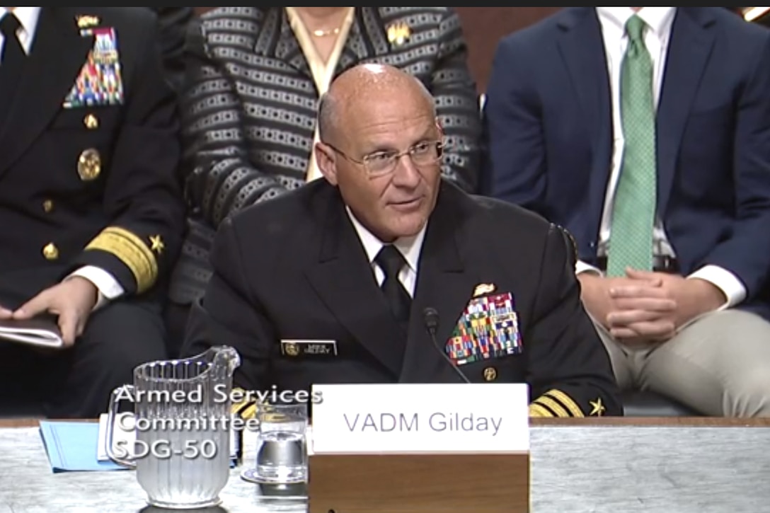 A Naval officer speaks while seated at a table..