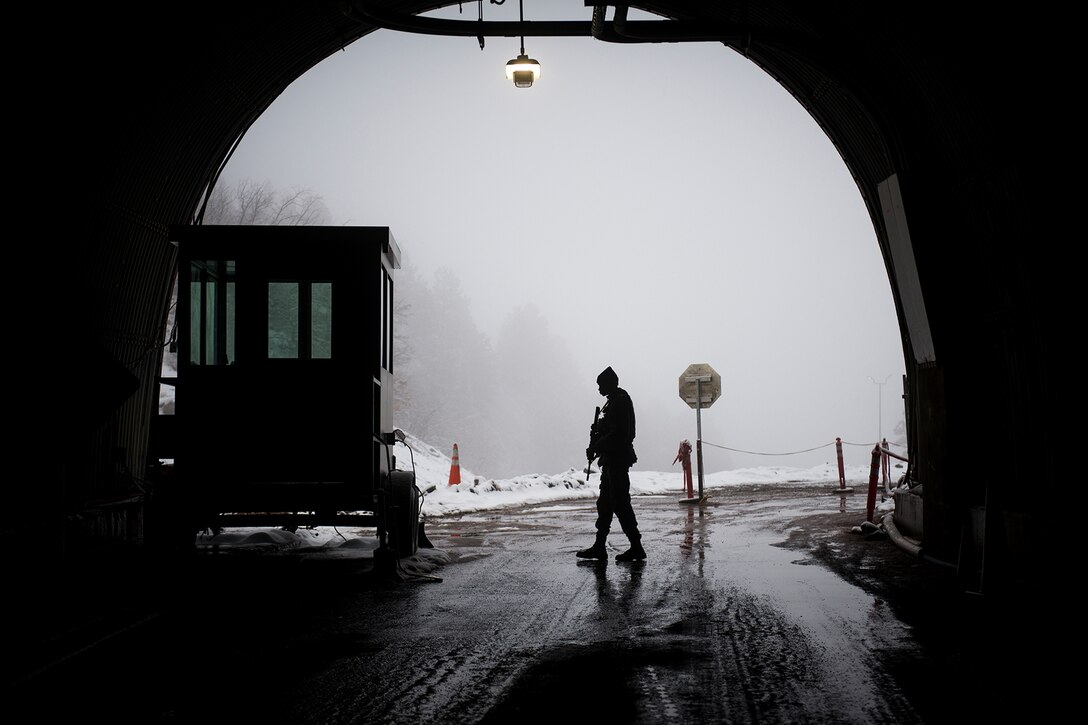 A service member, in silhouette, paces under an arch. He is carrying a weapon. Outside, snow is on the ground.