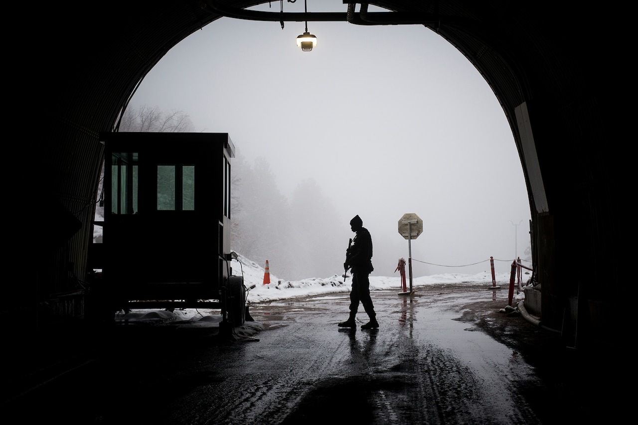 A service member, in silhouette, paces under an arch. He is carrying a weapon. Outside, snow is on the ground.