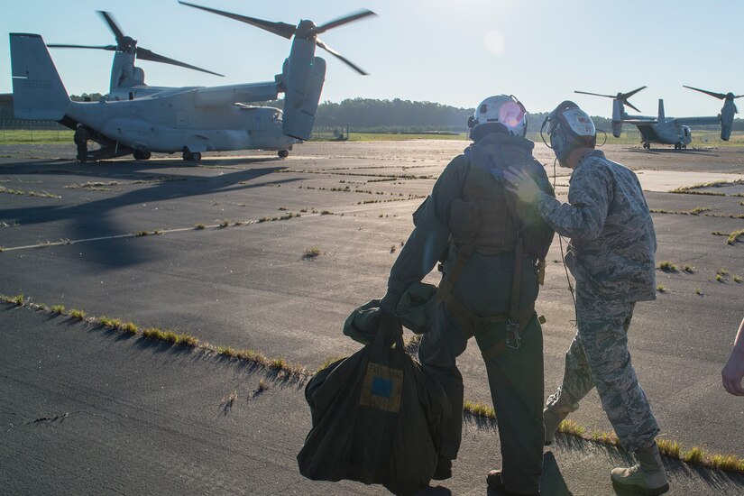 Two men walk across a concrete landing pad and approach a tilt-rotor aircraft.