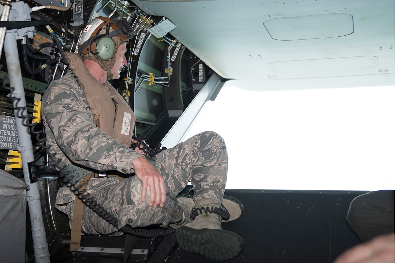 A man in a military uniform sits in the rear of an aircraft. He is wearing headphones. The tail of the aircraft is open.